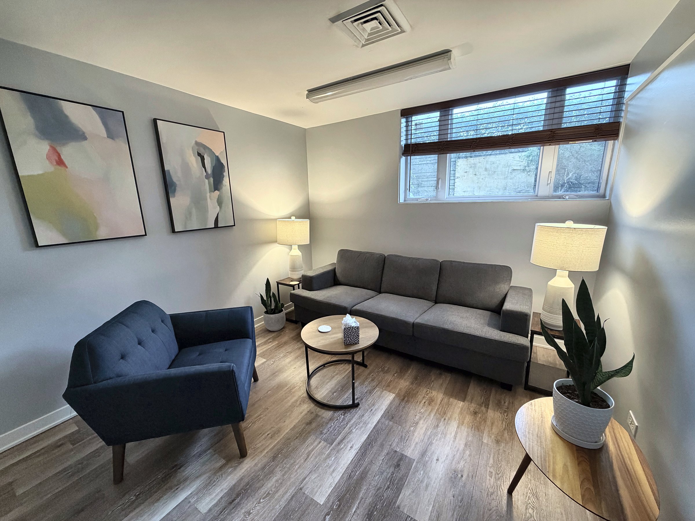 Evanston therapy room with a navy armchair, charcoal sofa, round wooden table, pastel wall art, and a warm lamp